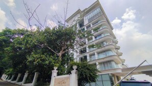 Ένα πολυώροφο κτίριο με μπαλκόνια, ένα δέντρο μπροστά και μια πινακίδα με κείμενο.