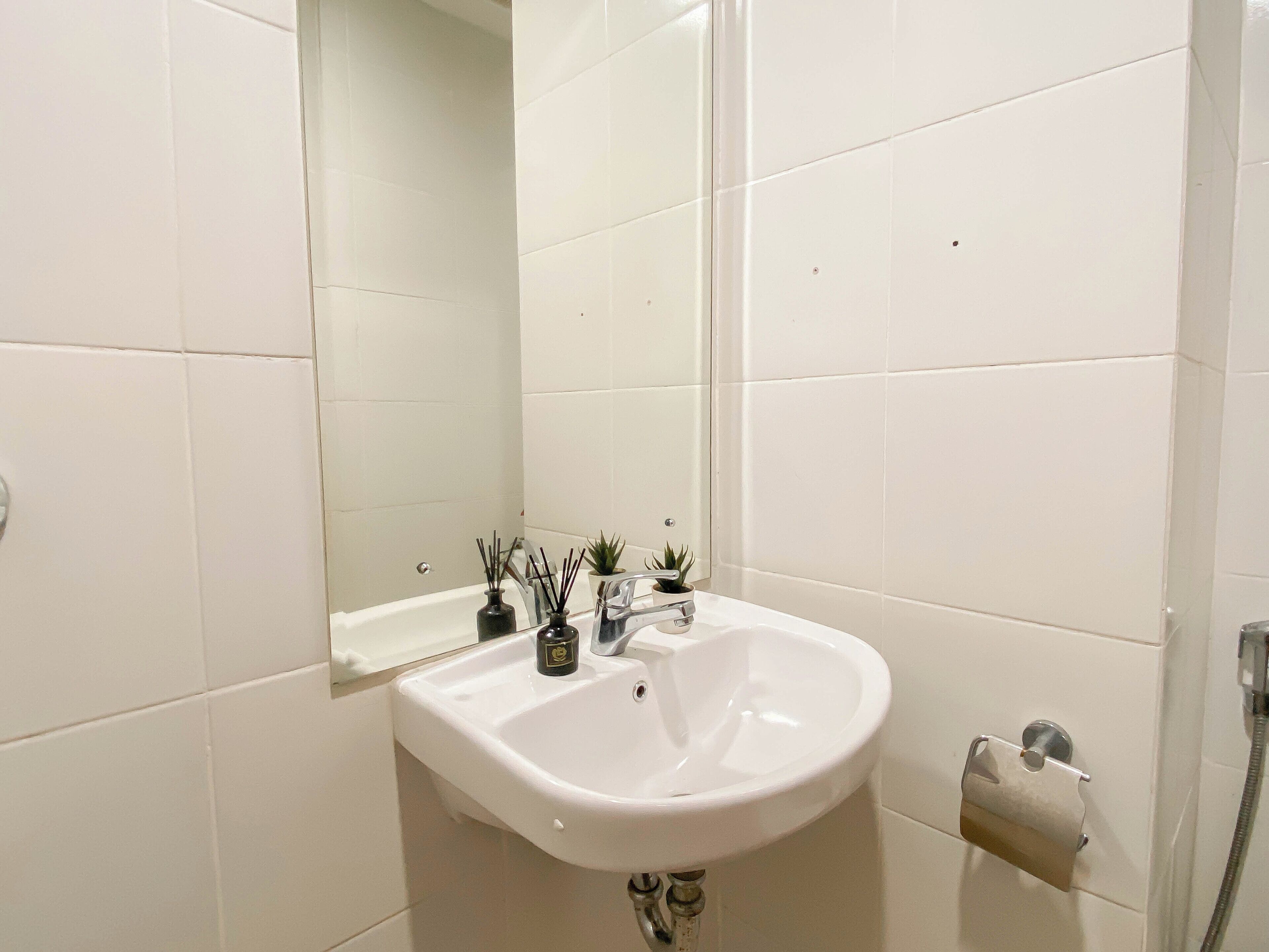 A bathroom with white tiles, a white sink, a mirror, a small potted plant, and a towel dispenser.