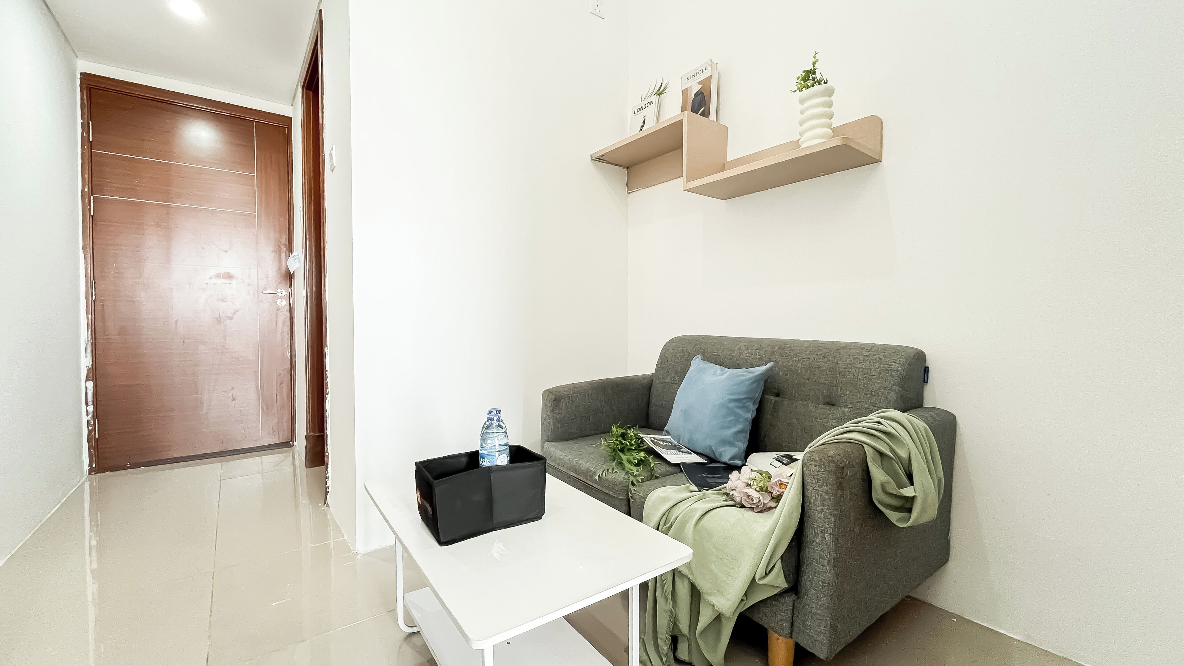 A modern living room with a grey sofa, a white coffee table, and a black storage unit. There is a wooden door in the background and a wall-mounted shelf with decorative items.