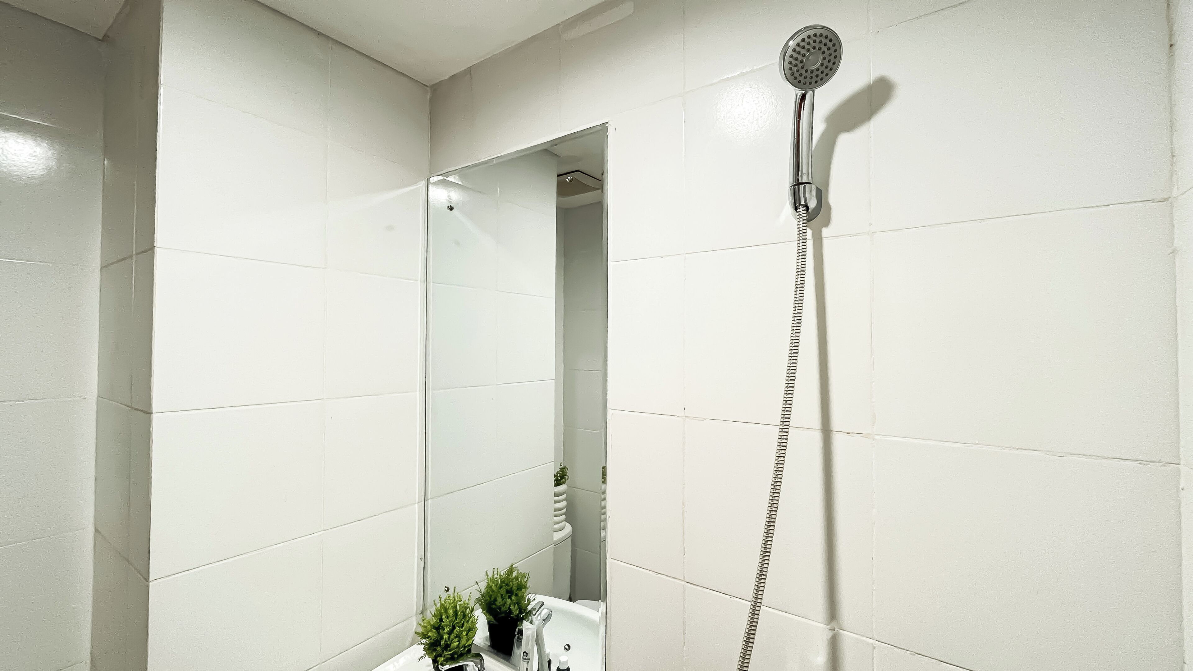 A bathroom with white tiles, a shower with a handheld showerhead, a sink with a mirror, and a small potted plant.