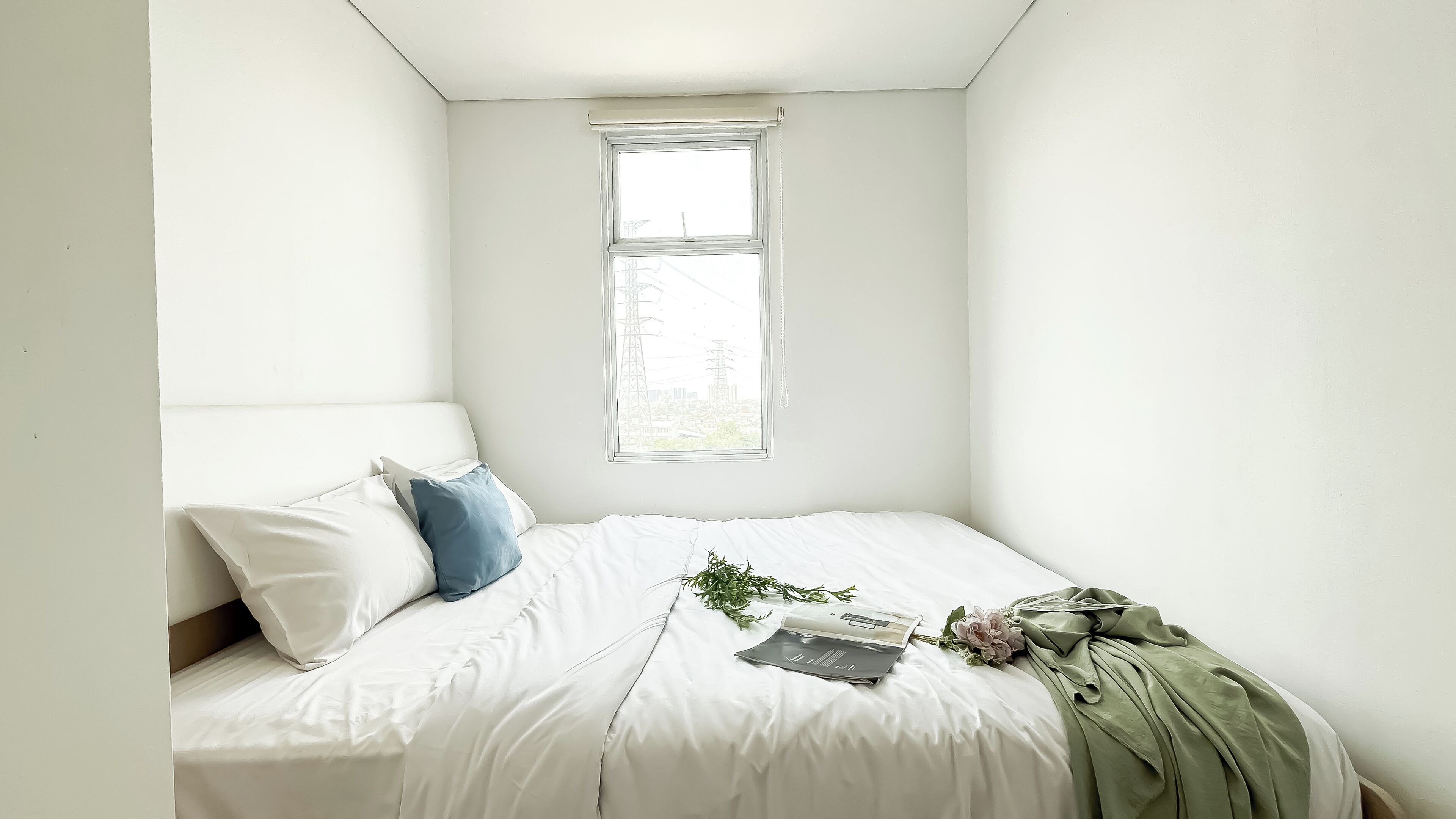 Een net opgemaakt bed met witte lakens en een blauw kussen, een raam met uitzicht op gebouwen, en een groene deken op het bed.