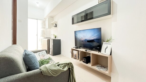 A modern living room with a grey sofa, a flat-screen TV mounted on the wall, a small kitchen area with a fridge and a kettle, and a shelf with books and a plant. - Restful 2Br At Vittoria Residence Apartment (Jakarta)