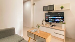 A modern living room with a flat-screen TV mounted on the wall, a wooden coffee table, a grey sofa, and a small potted plant. - Convenient 1Br Apartment At Vittoria Residence (Jakarta)