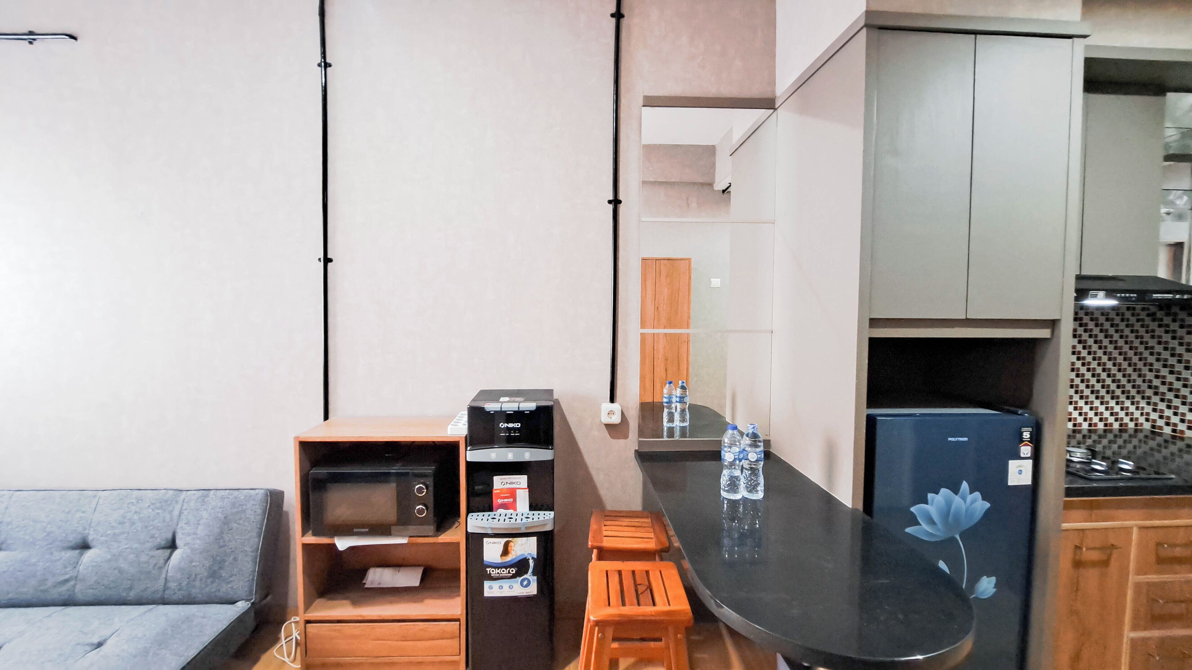A compact kitchen with a microwave, toaster, and water dispenser. A small wooden stool and a grey sofa are also visible.