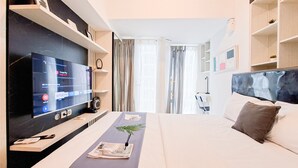 A modern hotel room with a large flat-screen TV, a bed with a headboard, a bedside table with a plant, and a window with curtains. - Minimalist And Restful Studio At Tokyo Riverside Pik 2 Apartment (Teluknaga)