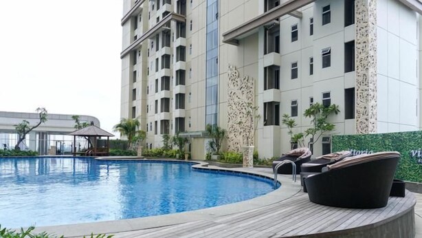 A swimming pool with a wooden deck, lounge chairs, and a building with multiple floors and balconies in the background. - Simply Look 1Br At Vittoria Residence Apartment (Jakarta)