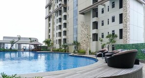 A swimming pool with a wooden deck, lounge chairs, and a building with multiple floors and balconies in the background. - Simply Look 1Br At Vittoria Residence Apartment (Jakarta)