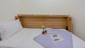 Ein Einzelbett mit Holz-Kopfstück, weißer Bettwäsche und einem kleinen Tisch mit Zeitung und Blumenvase.