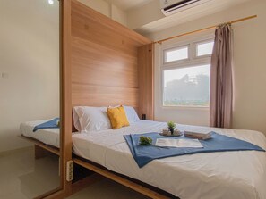 A bedroom with a wooden headboard, a single bed with white bedding and a blue throw, a window with curtains, and a small plant on the bed. - Good And Homey 2Br Apartment At Green Pramuka City (Jakarta)