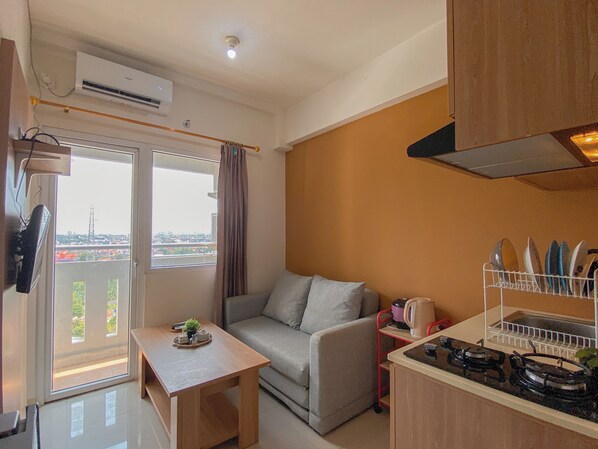 A compact living space with a sofa, a small table, a kitchen area with a gas stove, and a view through a large window. - Good And Homey 2Br Apartment At Green Pramuka City (Jakarta)