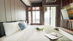 A modern bedroom with a large bed, a view of the city through the window, and a mounted TV on the wall. - Cozy Stay Studio Apartment At M-Town Residence (Gading Serpong)
