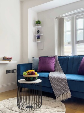 A modern living room with a blue sofa, a round coffee table with a fruit bowl, a white wall with shelves, and a window with curtains. - Elegant Shoreditch Apartment with Private Balcony (Greater London)