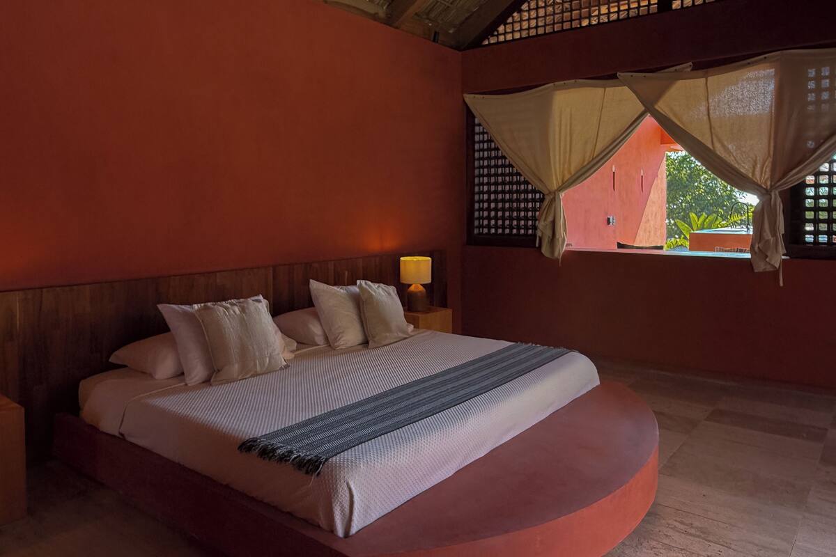 A bed with a curved wooden base, a headboard, and multiple pillows. There is a nightstand with a lamp and a window with a mosquito net.