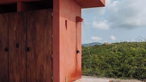 Ein Gebäude mit roter Fassade und Holztür, ein Kaktus im Vordergrund und eine bewachsene Hügelkulisse im Hintergrund.
