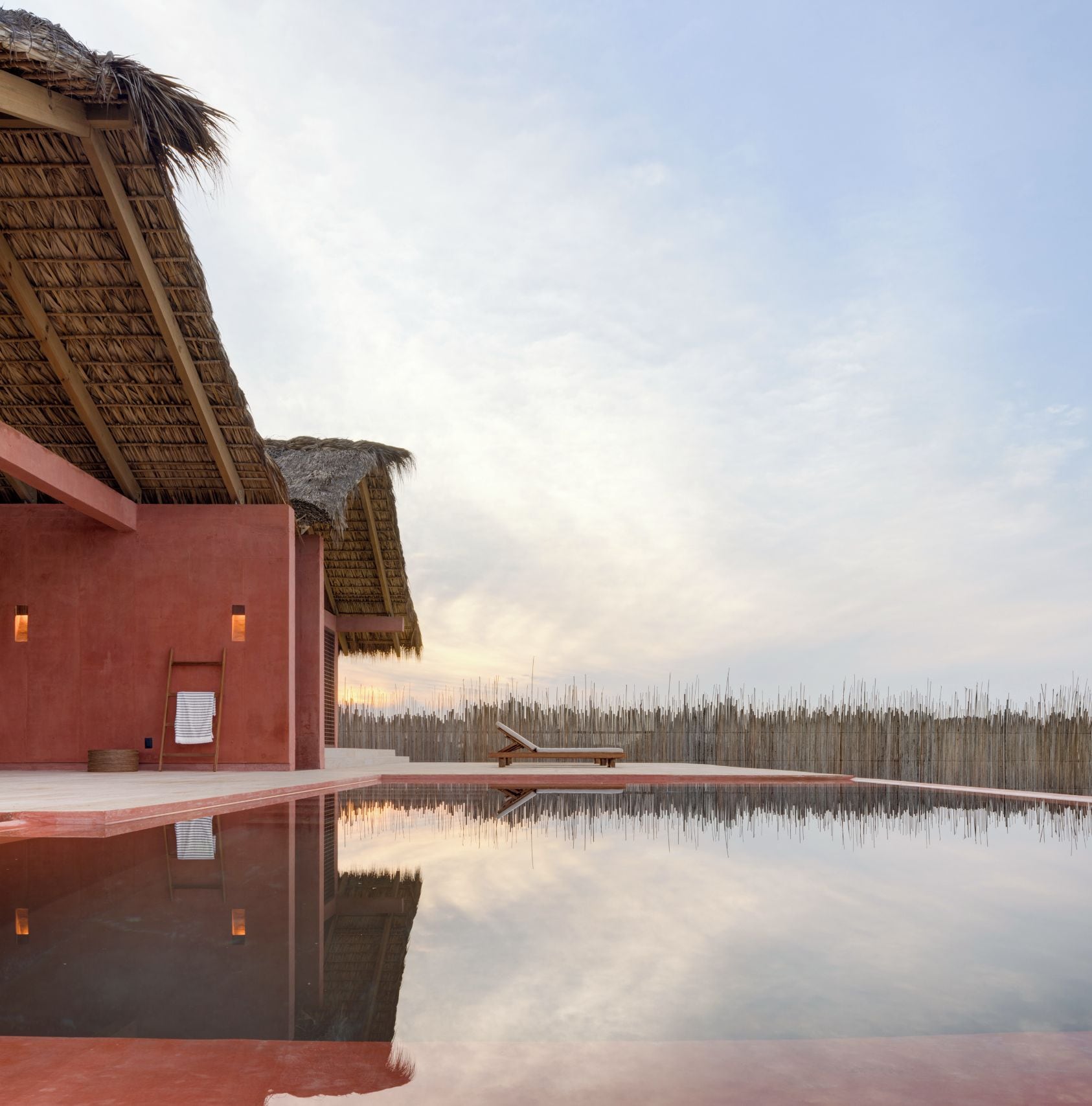 A serene pool area with a thatched-roof building, clear sky, and calm water reflecting the surroundings.
