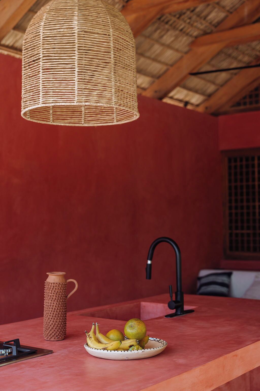 Une cuisine avec un luminaire tressé, un panier de fruits, un pichet en céramique et un robinet noir.