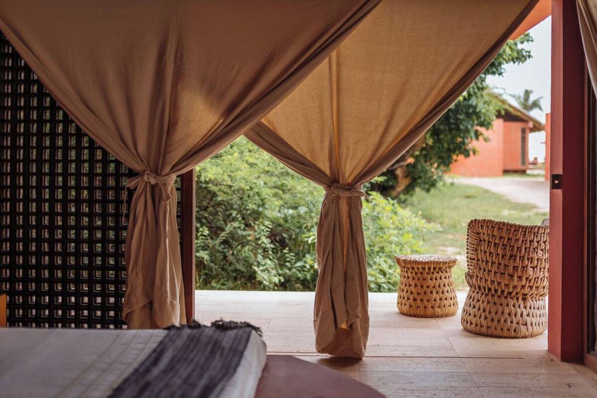 A room with a bed, a woven chair, and a view of greenery outside.