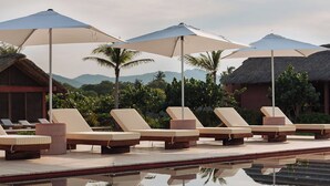 Un área junto a la piscina con sillones de descanso y sombrillas, rodeada de edificios con techo de paja y palmeras.