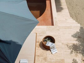 Un espacio al aire libre iluminado por el sol con piscina, un banco de madera y una cesta de mimbre con una toalla.