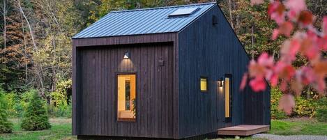 A small, dark wooden cabin with a sloped roof, a single visible door, and a small porch.