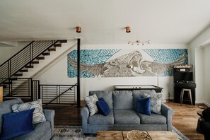 A living room with a staircase, a sofa set, a wall mural, and a piano. - Modern 3BR Condo with Rooftop View Near Deep Ellum & Fair Park (Dallas)