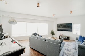 A spacious living room with a large window, a grey sofa, a coffee table, a pool table, and a flat-screen TV mounted on the wall.