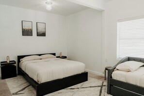 A bedroom with a bed, bedside tables, a nightstand, a chair, a window with blinds, and two wall-mounted pictures. - Modern 3BR Condo with Rooftop View Near Deep Ellum & Fair Park (Dallas)