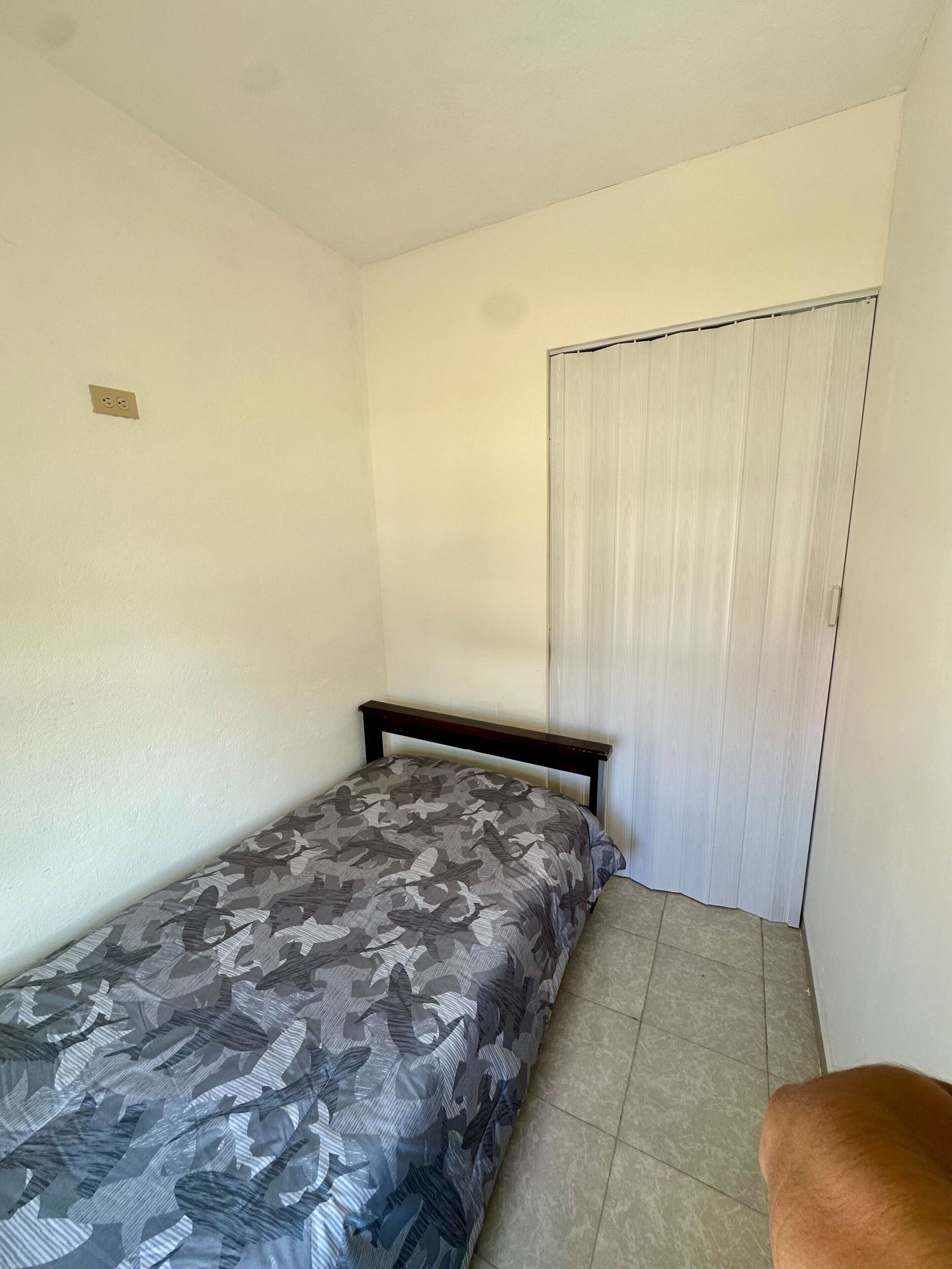Ein kleines Zimmer mit einem Einzelbett, einer gestreiften Bettdecke, einem Holz-Kopfteil, einem Fliesenboden und einer weißen Tür mit Vorhang.
