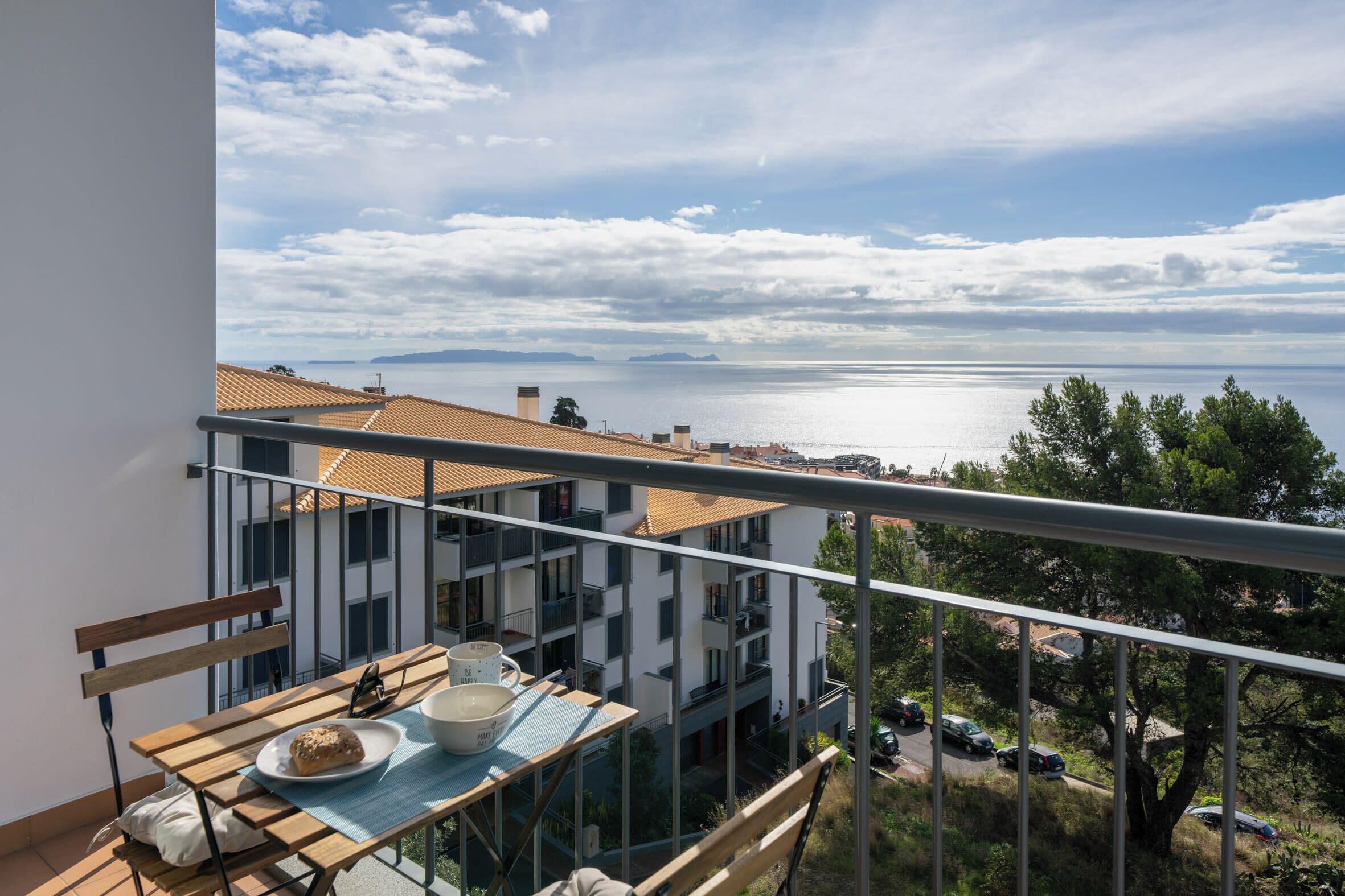 A balcony with a view of the sea, a table with a plate and a bowl, and a building with a tiled roof in the background.