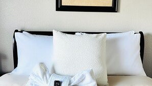 Ein ordentlich bezogenes Bett mit weißen Leinen, einer Fernbedienung und einem Schleifen-Kissen. Über dem Bett hängt ein gerahmtes Blumenbild.