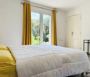 A bedroom with a bed, a window with curtains, a door, and a radiator. - L'Enclos des Chênes Verts (VENTENAC CABARDES)