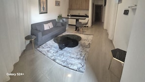 Un salon moderne avec un canapé gris, une table basse et un tapis. On aperçoit au fond une cuisine avec des meubles en bois et un coin repas.