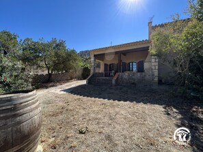 A stone house with a covered porch, a stone wall, and a dry, leaf-covered ground.