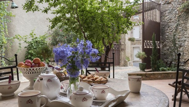 A round table with a tray holding a teapot, cups, a bowl of fruit, and a vase with flowers. - Aude à la Vigne - CRUSCADES (CRUSCADES)