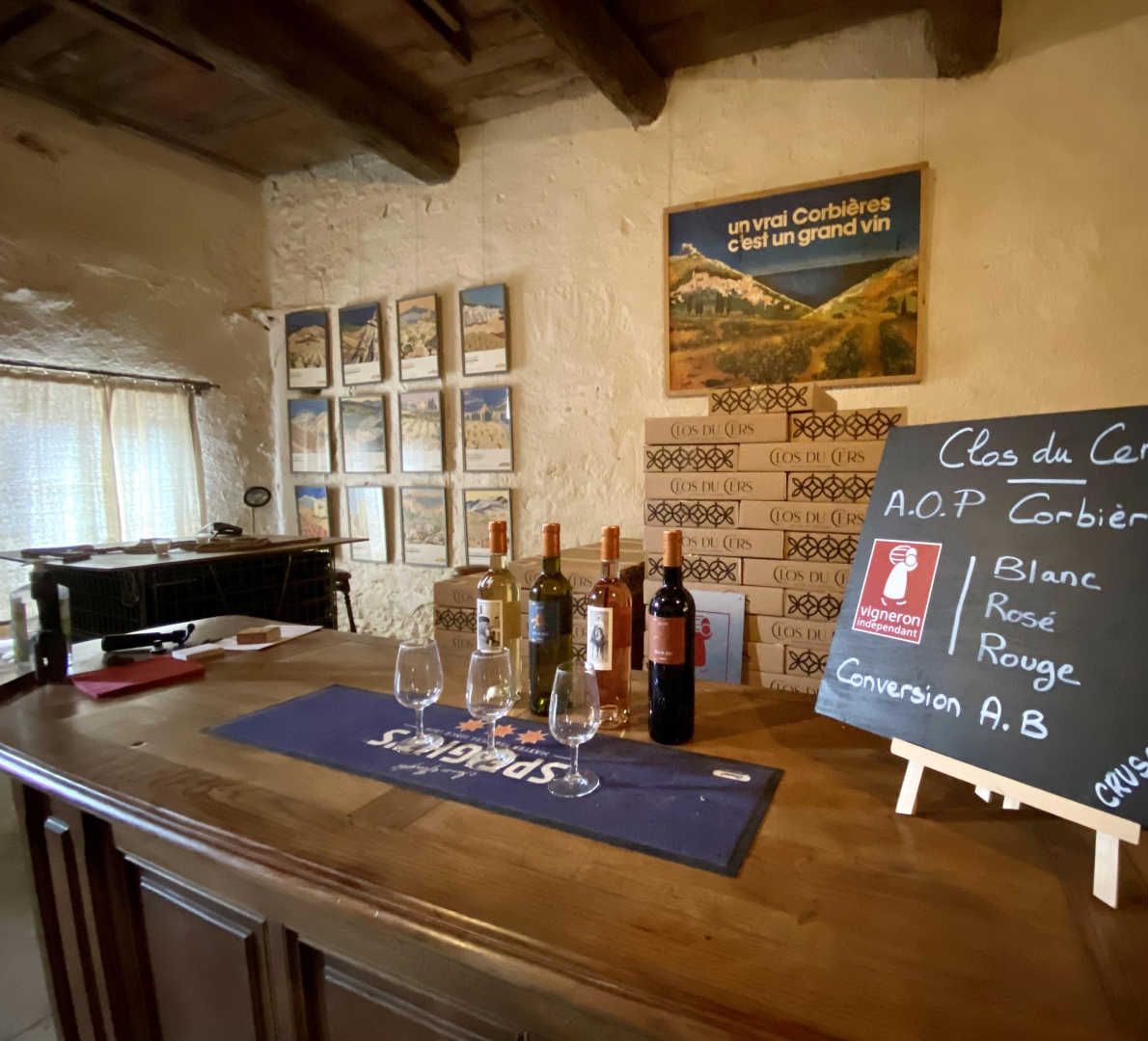 Un espace dédié à la dégustation de vins, avec un comptoir en bois, des bouteilles de vin, des verres et un tableau noir affichant la carte des vins proposés.