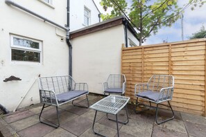 Un patio con sillas y una mesa de metal, una valla de madera y un edificio blanco con una ventana.