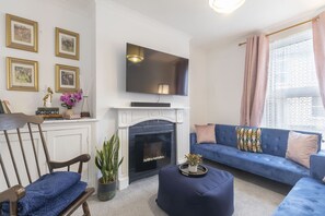A living room with a fireplace, a blue sofa, a television mounted above the fireplace, and framed pictures on the wall. - Central 3 Bedroom Home for 7 Guests (Cheltenham)