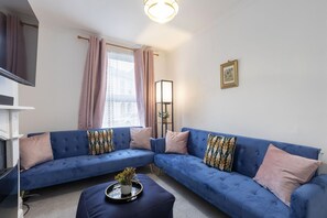 A living room with a blue sectional sofa, a small coffee table with a plant, a floor lamp, and a picture on the wall. - Central 3 Bedroom Home for 7 Guests (Cheltenham)