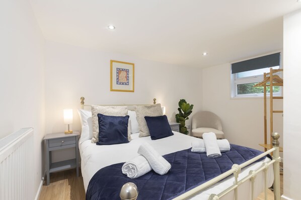A neatly made bed with a blue quilt, white linens, and rolled towels. There is a bedside table with a lamp, a framed picture on the wall, and a window with a view of greenery.