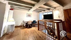 Una habitación rústica con vigas de madera a la vista, una zona de comedor con sillas y una mesa, un televisor y una escalera.