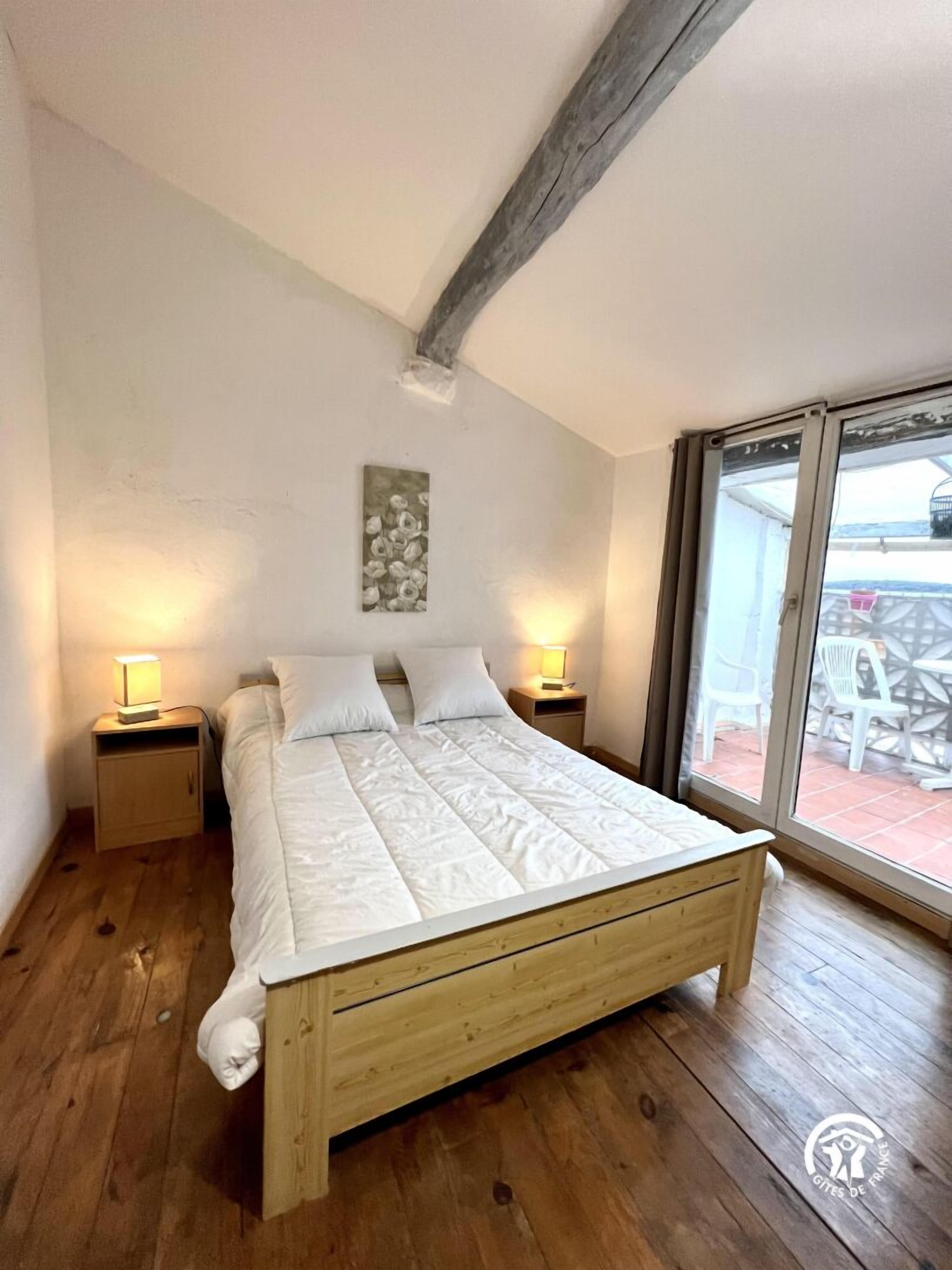 Un dormitorio con una cama de madera, dos mesitas de noche con lámparas, un techo con vigas a la vista y vistas a muebles de exterior a través de un ventanal.
