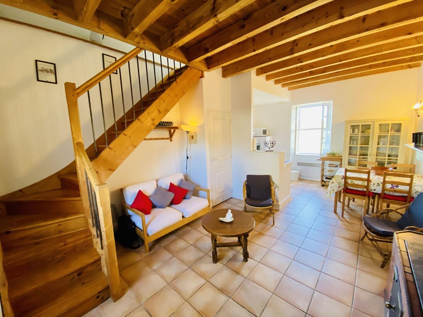 A cozy interior with a wooden staircase, a sofa with cushions, a dining table with chairs, and a kitchen area.