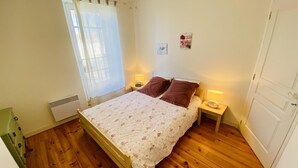 A bedroom with a wooden bed, a radiator, a window with curtains, and a small bedside table with a lamp. - Gite du Cammas Blanc (VILLEPINTE)