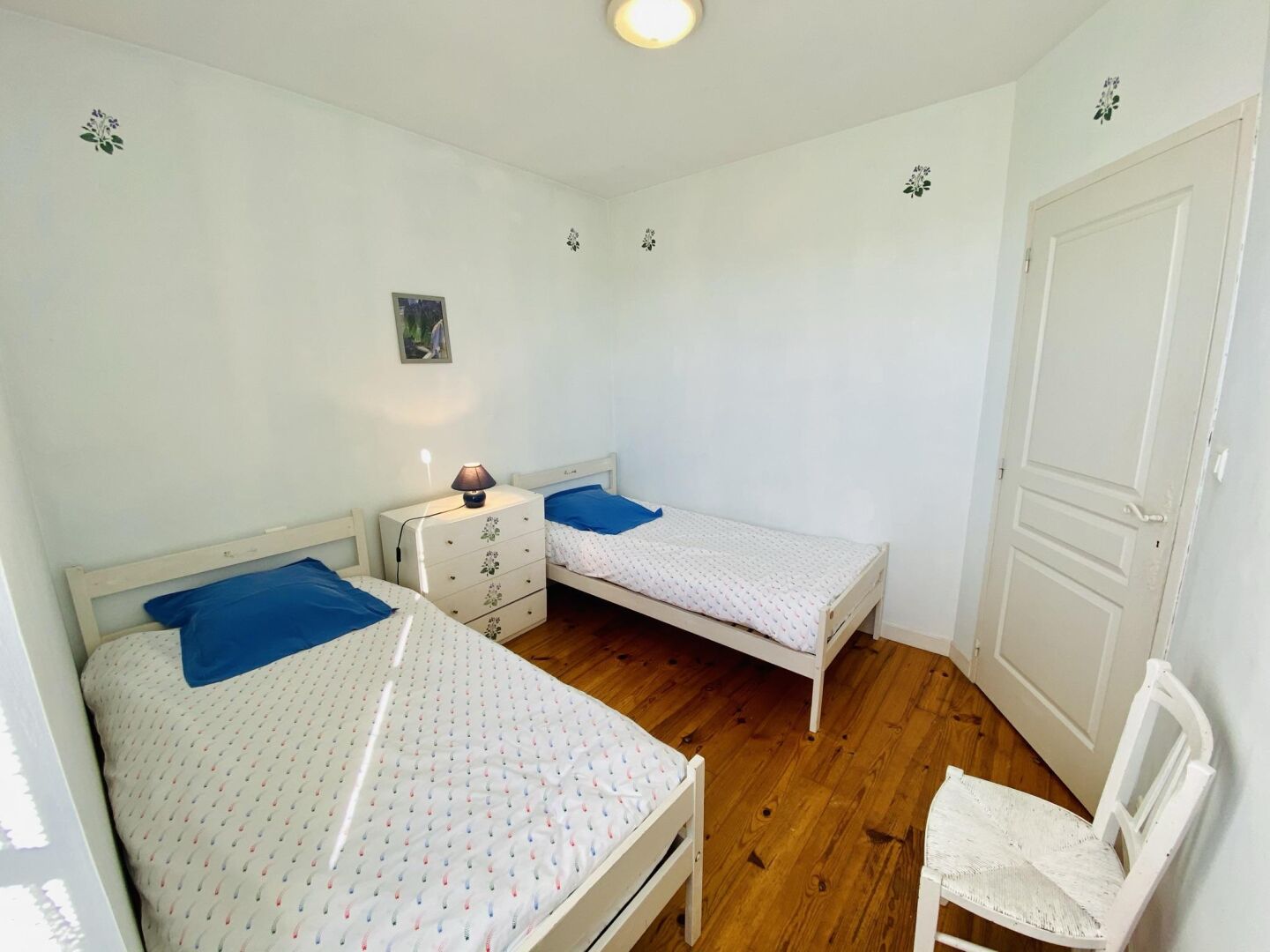 A small, clean room with two single beds, a nightstand, a chair, and a door.