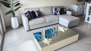 A modern living room with a grey sectional sofa, a glass coffee table with a blue marble design, and a large potted plant.