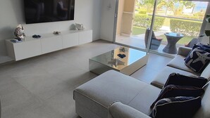 A modern living room with a flat-screen TV, a sofa, a coffee table, and a view of a garden and pool.