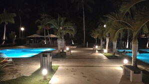 En nattbild av ett semesterområde med en swimmingpool, palmer och en väl upplyst gångväg.