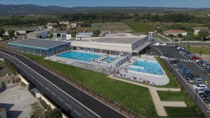 Un complexe de piscines comprenant plusieurs bassins et une zone couverte, situé à proximité d’une route et d’un parking.