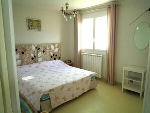 Een slaapkamer met een bed, een radiator, een raam met gordijnen, een kleine tafel en een kroonluchter.