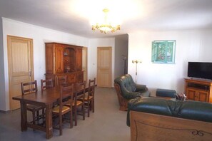 Een eetgedeelte met een houten tafel en stoelen, een groene leren bank, een televisie en een kroonluchter.
